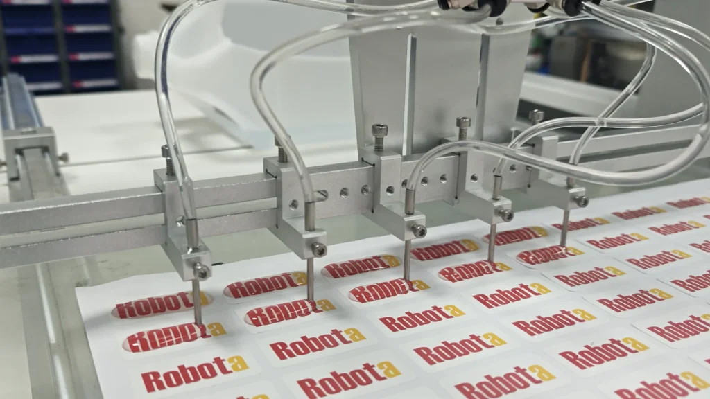 Multiple dispensing nozzles applying resin to regularly arranged Robota logo sticker sheets